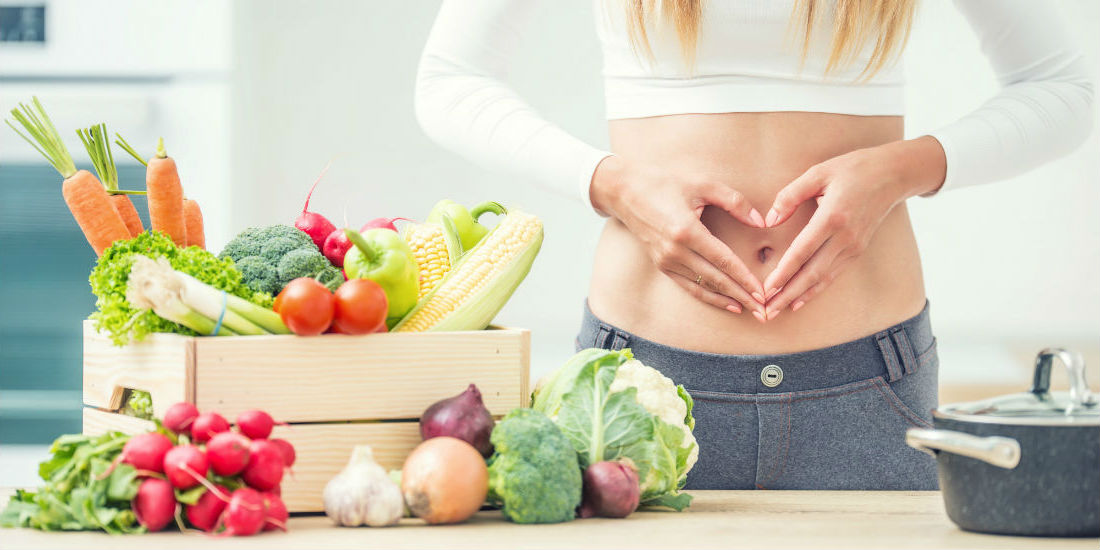 Alimentazione e fertilità: una ragazza con le mani a cuore sulla pancia e intorno a lei frutta e verdura